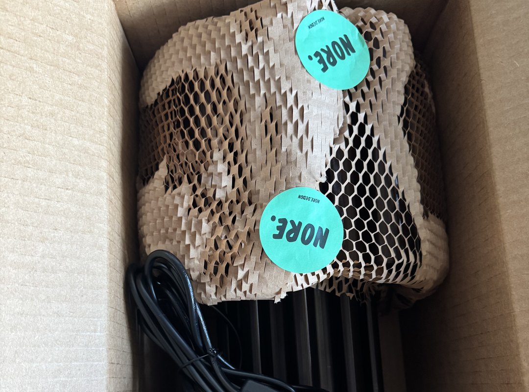 A lamp wrapped in paper honeycomb with two NORE stickers, packed in its shipping box with the cord